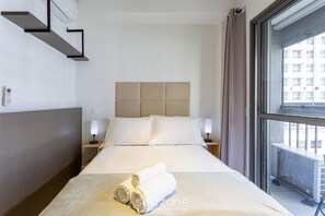 A hotel room with a bed, two bedside tables, a headboard, and a window with a view of buildings. - Complete Studio 350m From the Metro Thk0502 (São Paulo)