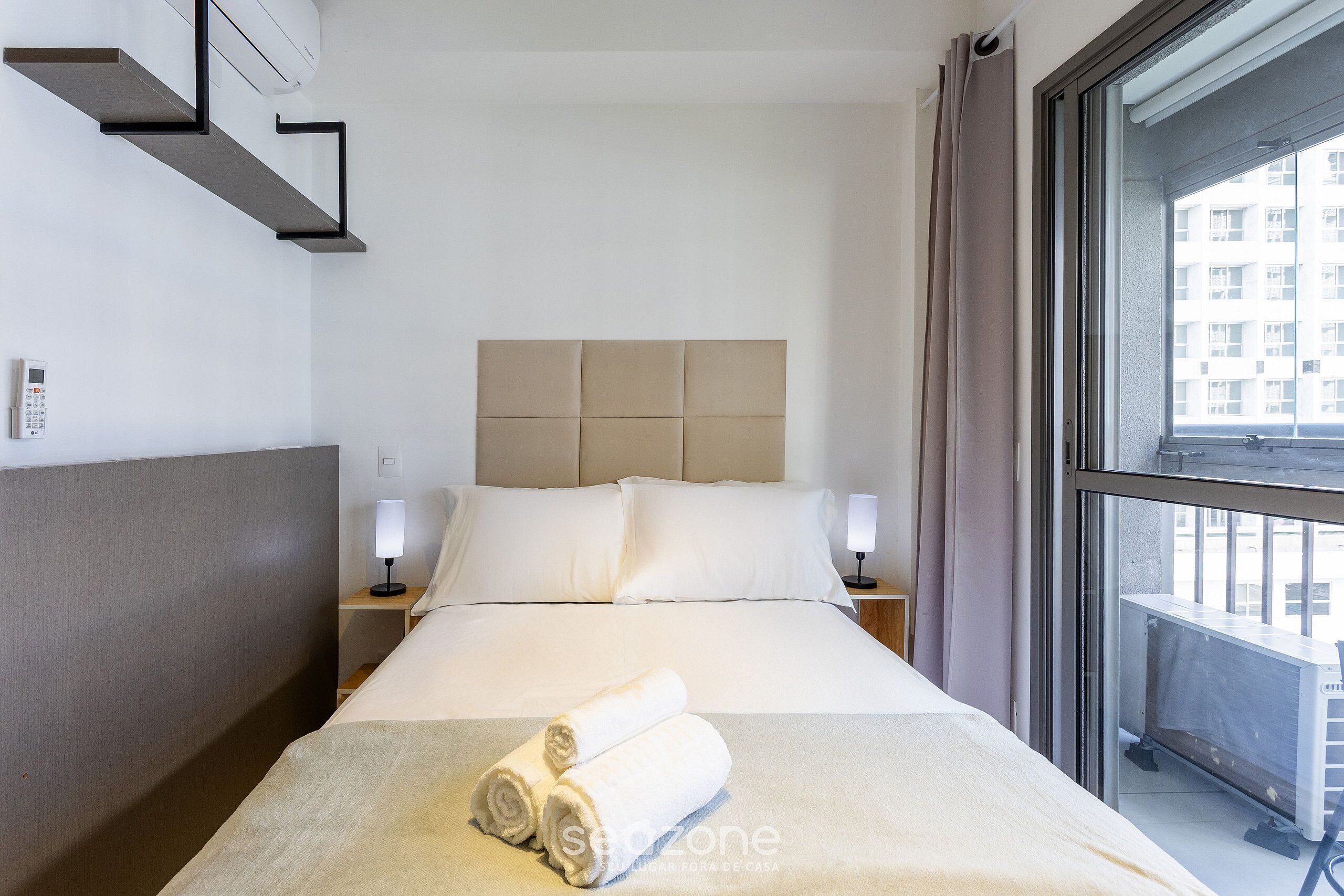 A hotel room with a bed, two bedside tables, a headboard, and a window with a view of buildings.