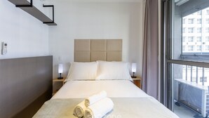 A hotel room with a bed, two bedside tables, a headboard, and a window with a view of buildings. - Complete Studio 350m From the Metro Thk0502 (São Paulo)