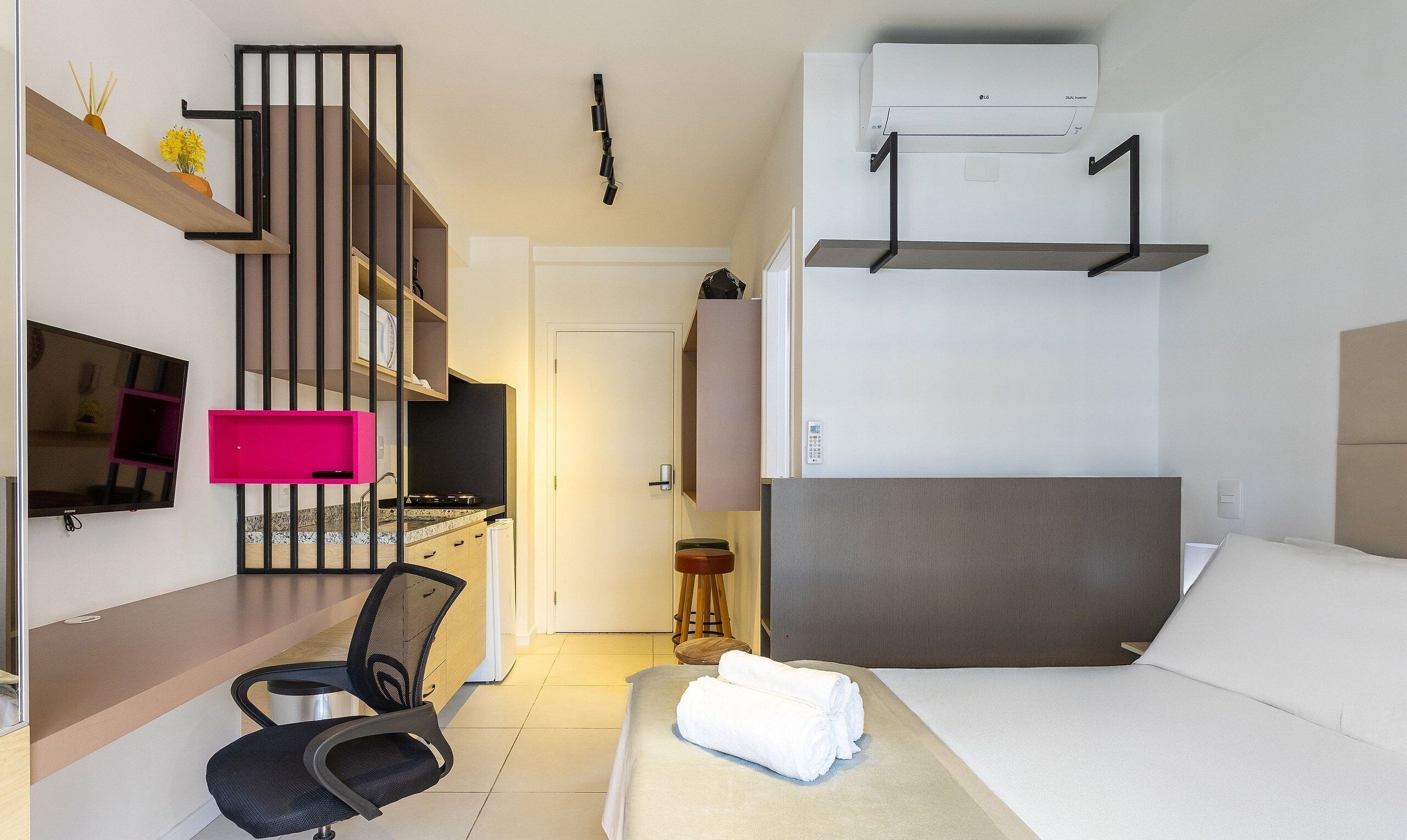 Quarto de hotel compacto com cama, escrivaninha com cadeira, televisão, cozinha compacta e banheiro.