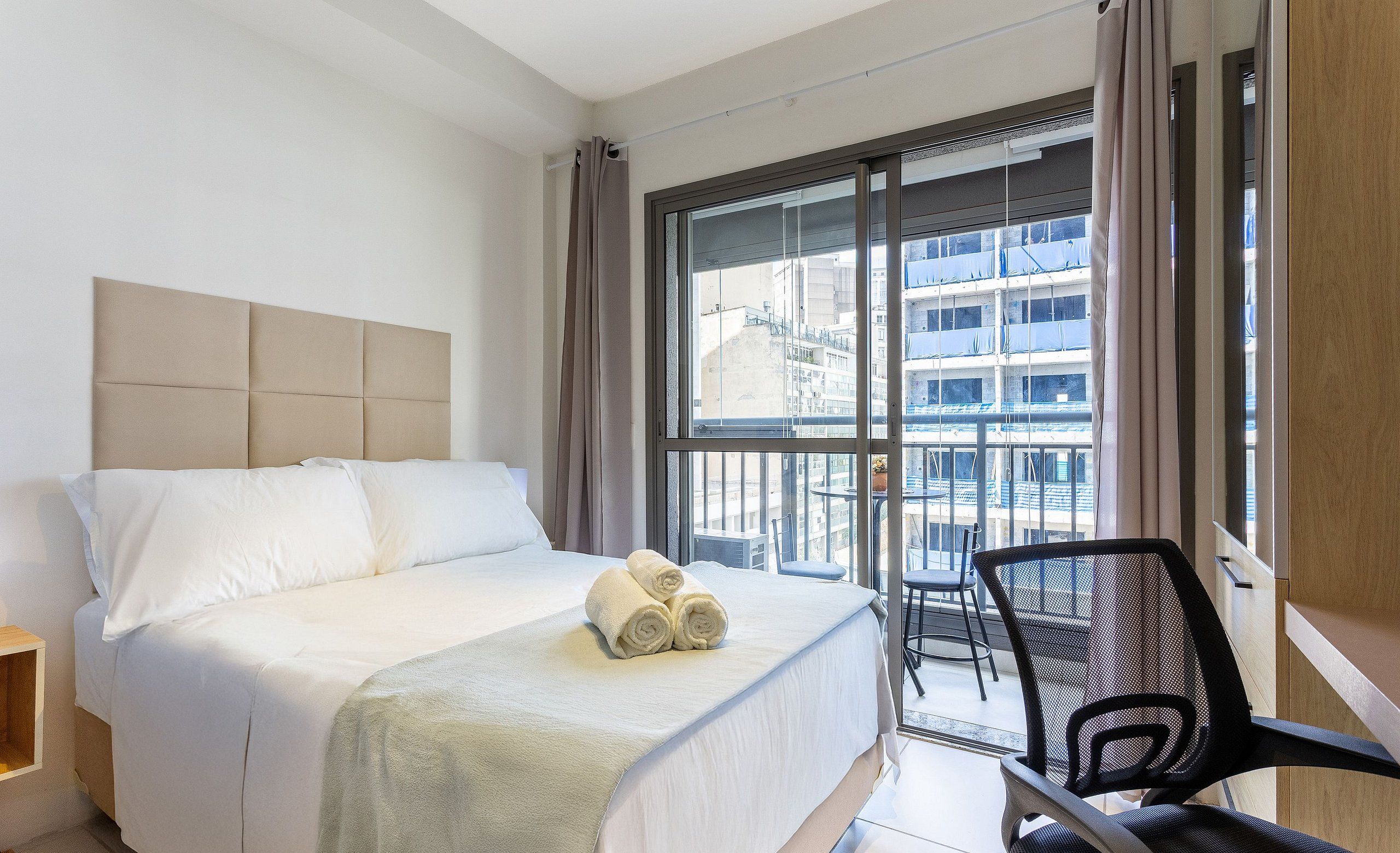 Een hotelkamer met een bed, een stoel en een groot raam met uitzicht op gebouwen.