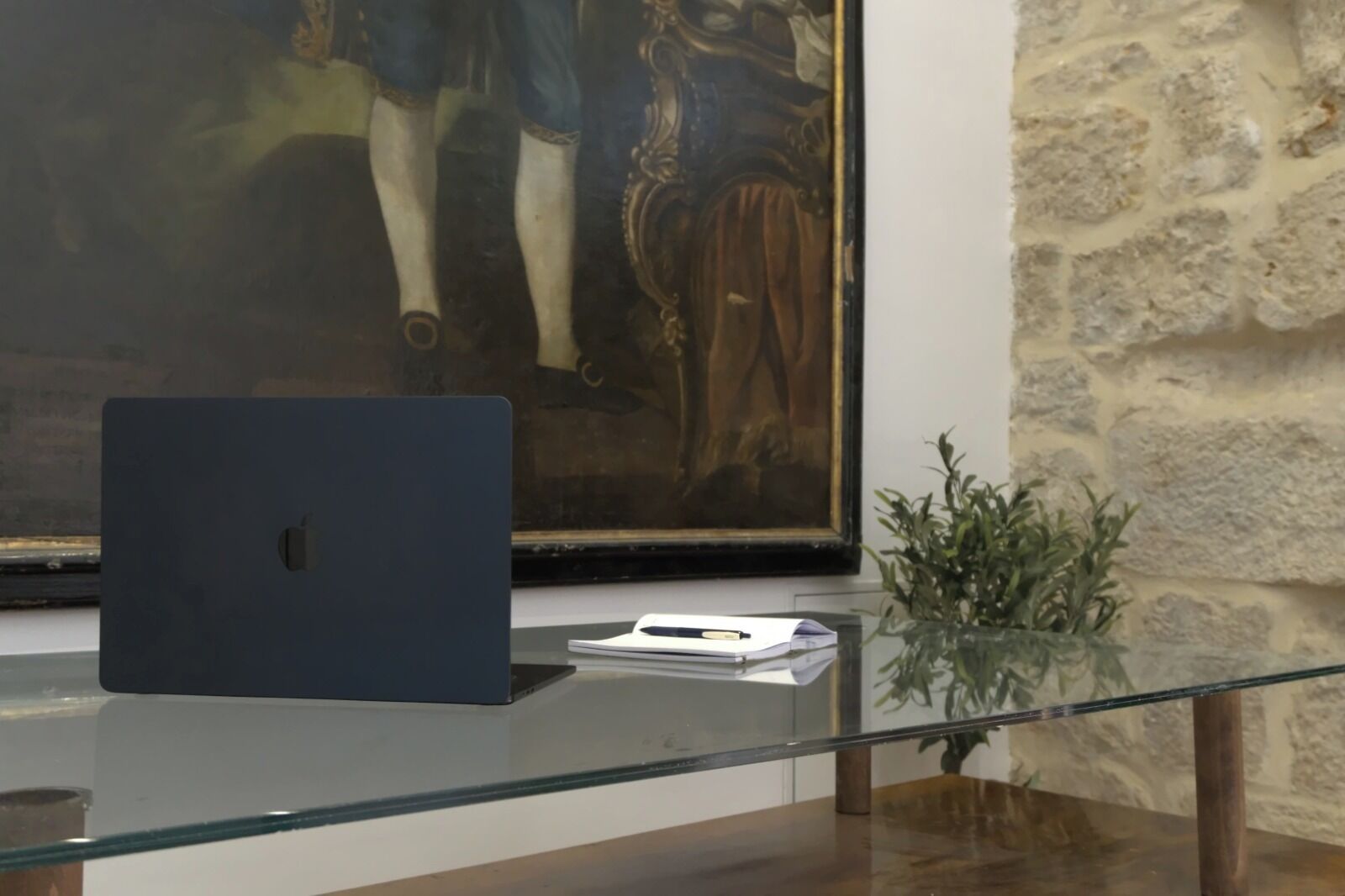 A laptop on a glass table with a framed painting in the background.