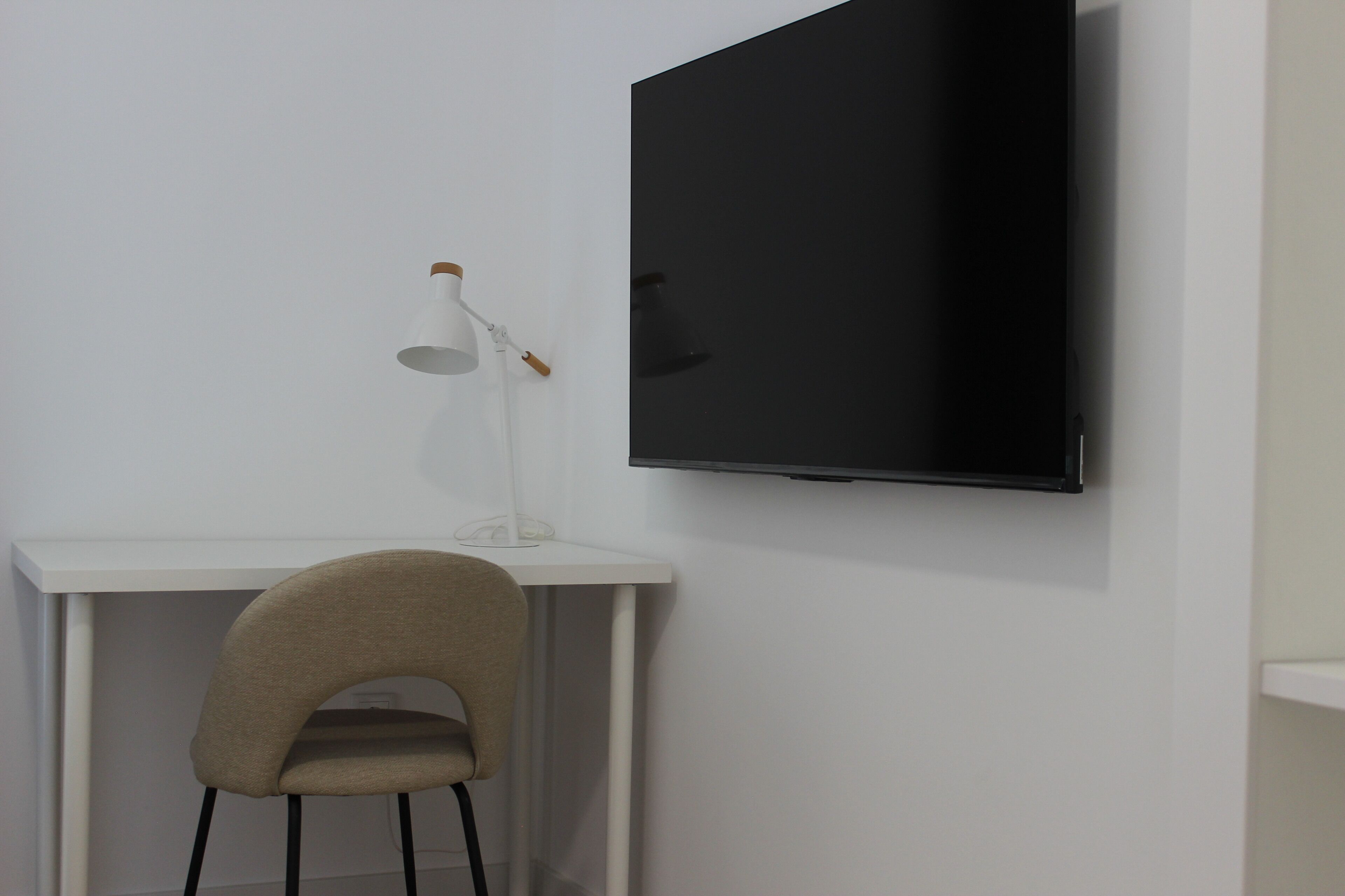 Et værelse med et hvidt skrivebord, en stol og et fladskærms-tv monteret på væggen.