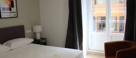 A hotel room with a bed, a chair, a nightstand, a lamp, and a window with curtains.