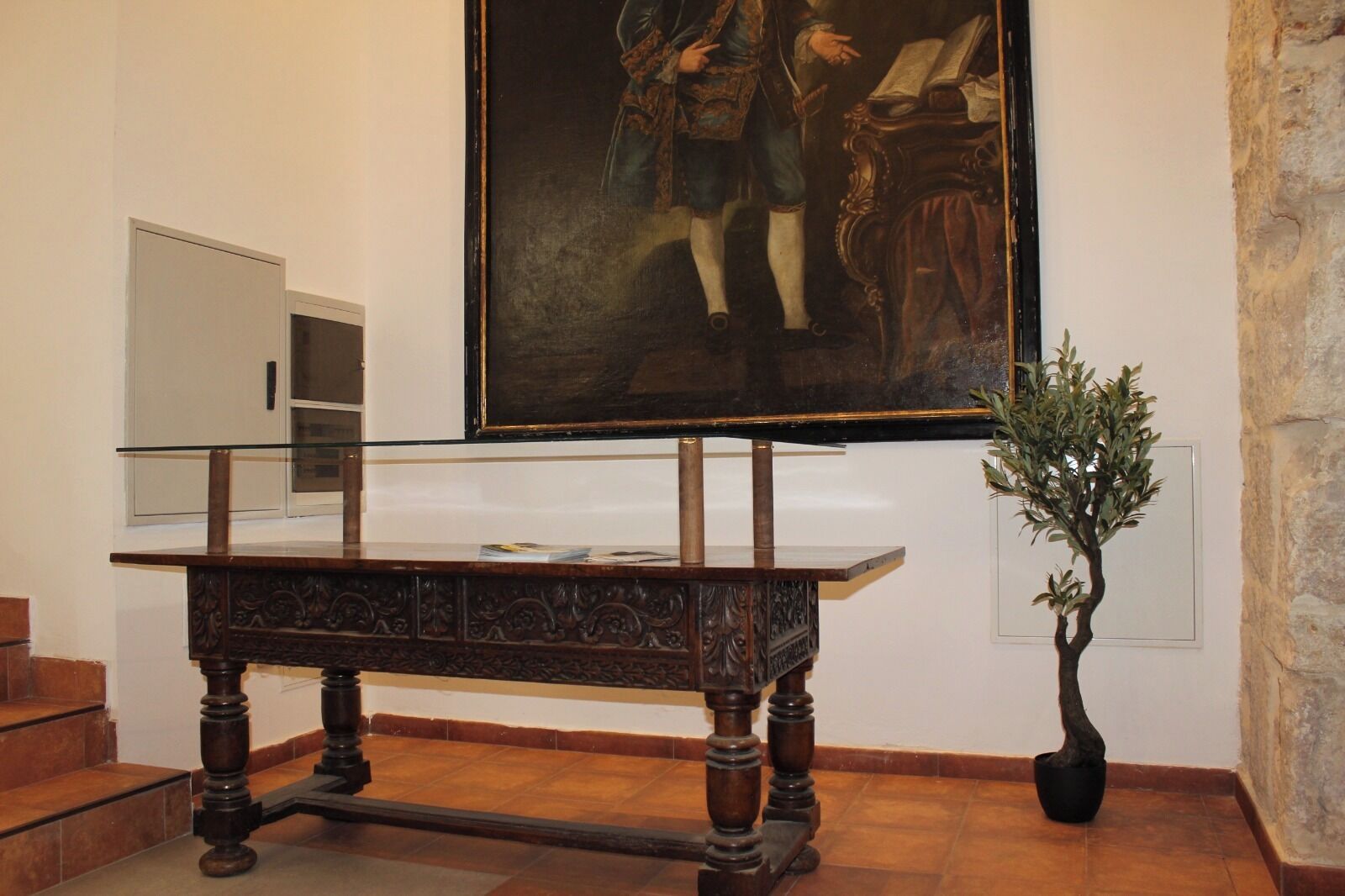 Une pièce avec un grand tableau, une table en bois surmontée d’une vitre, et une plante en pot.