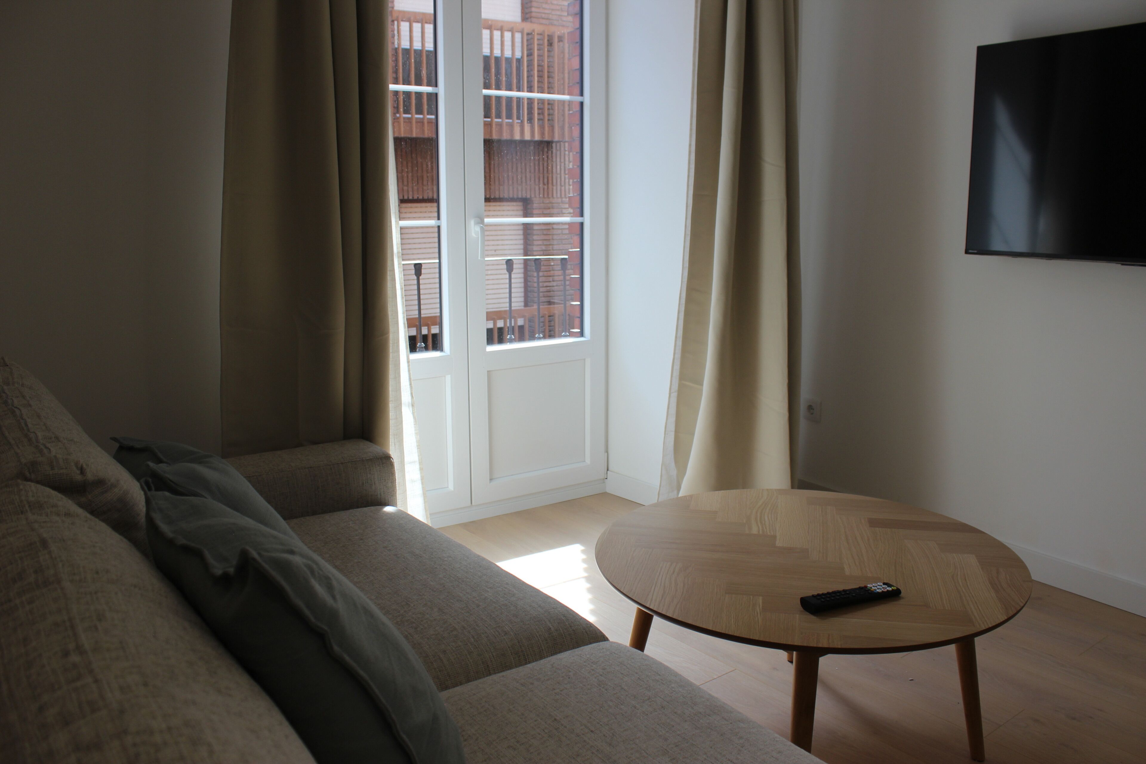 Una sala de estar con un sofá, una mesa de centro redonda de madera, un televisor montado en la pared y un puertas correderas de vidrio que dan a un balcón al aire libre.