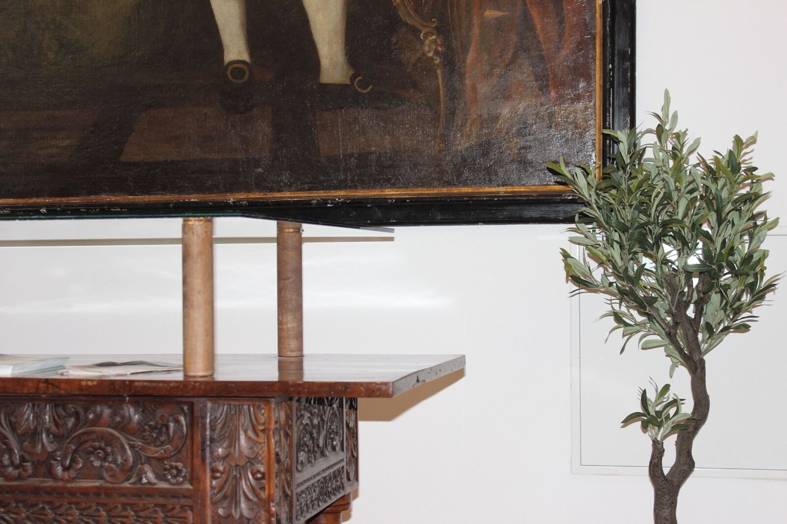 A wooden table with a carved design, a framed painting above it, and a potted plant to the right.