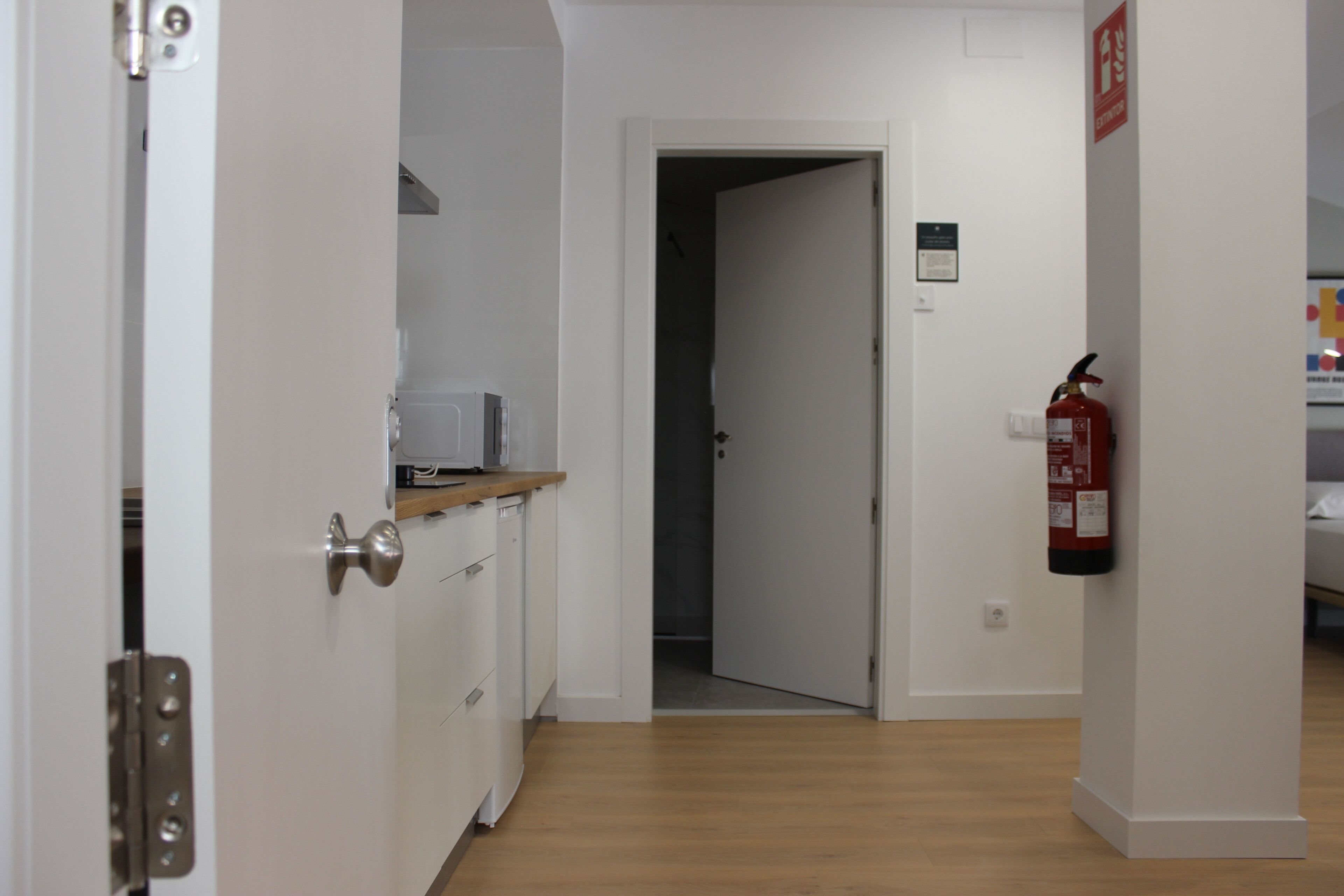 Corredor com área de cozinha à esquerda, uma porta que dá para outro quarto e um extintor de incêndio fixado na parede.
