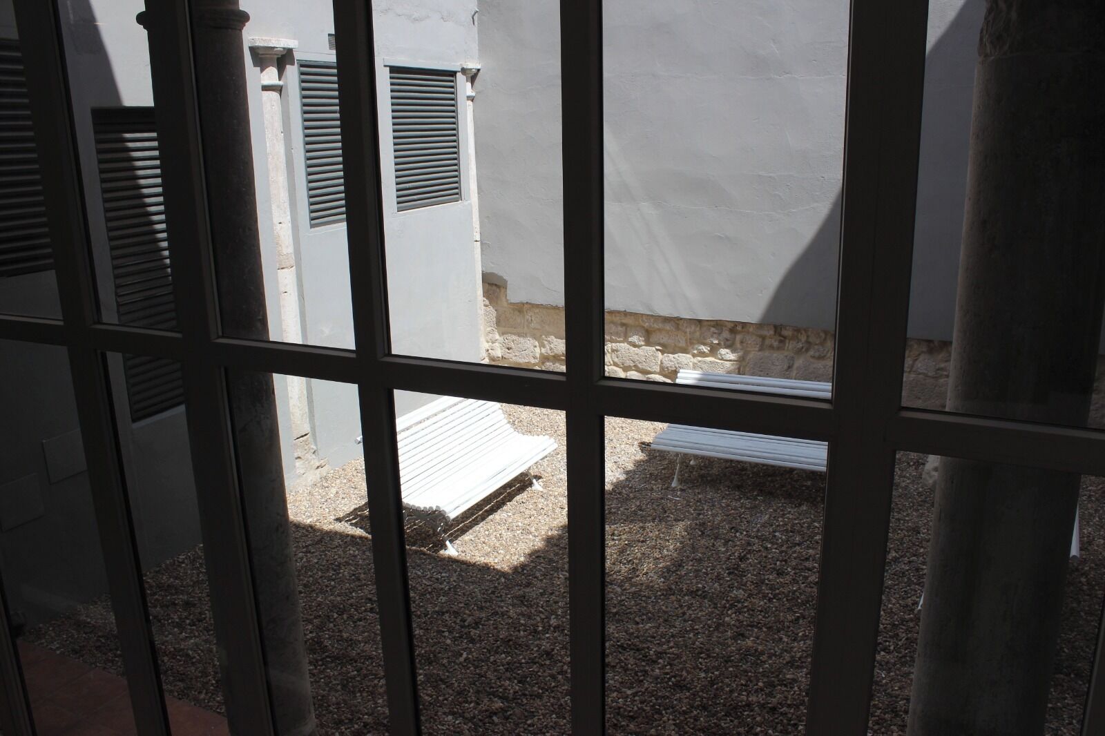 Vista a través de barrotes, mostrando un patio con un banco, un muro de piedra y un edificio con contraventanas.