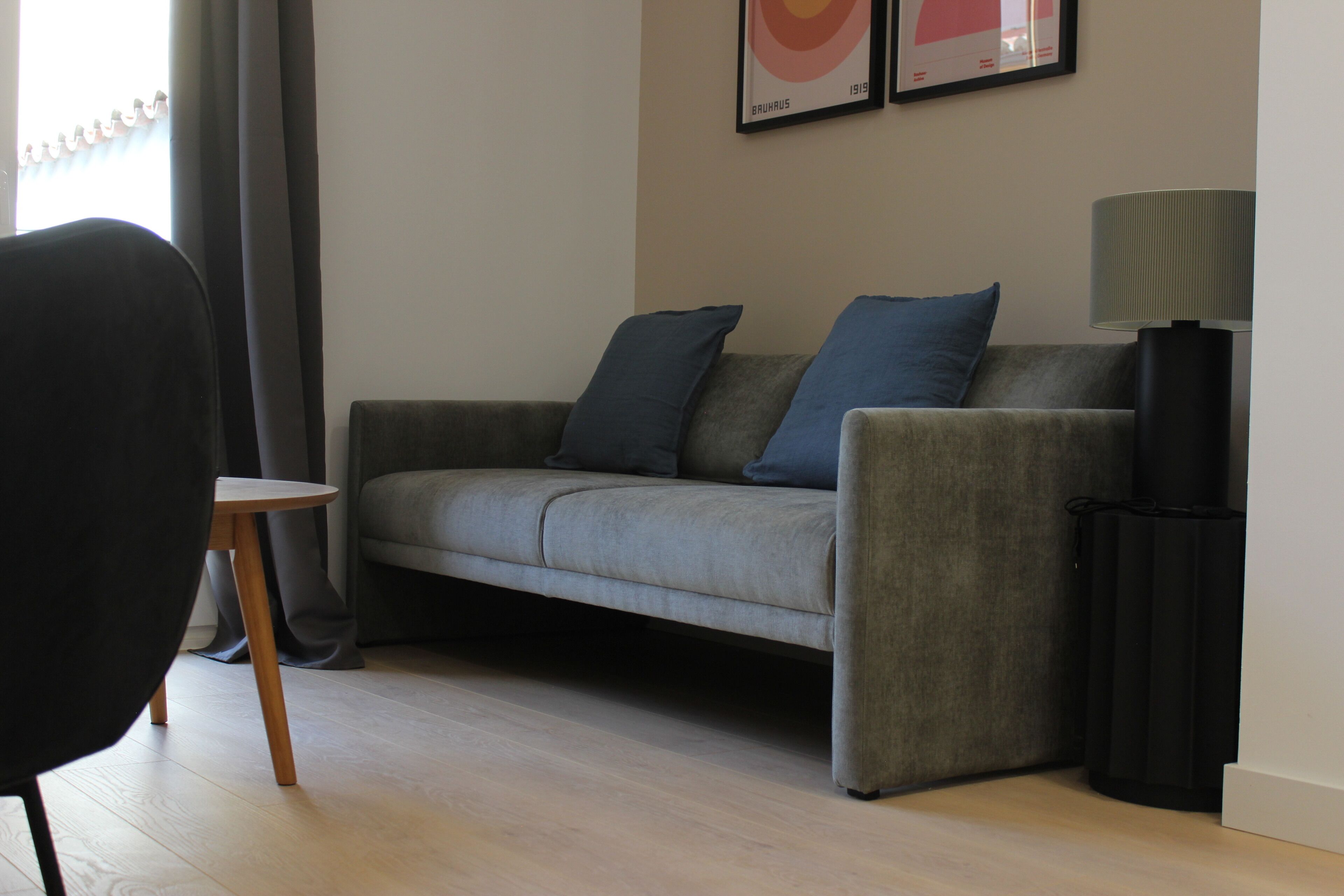 Een moderne woonkamer met een grijze bank, twee blauwe kussens, een kleine ronde tafel en een vloerlamp.