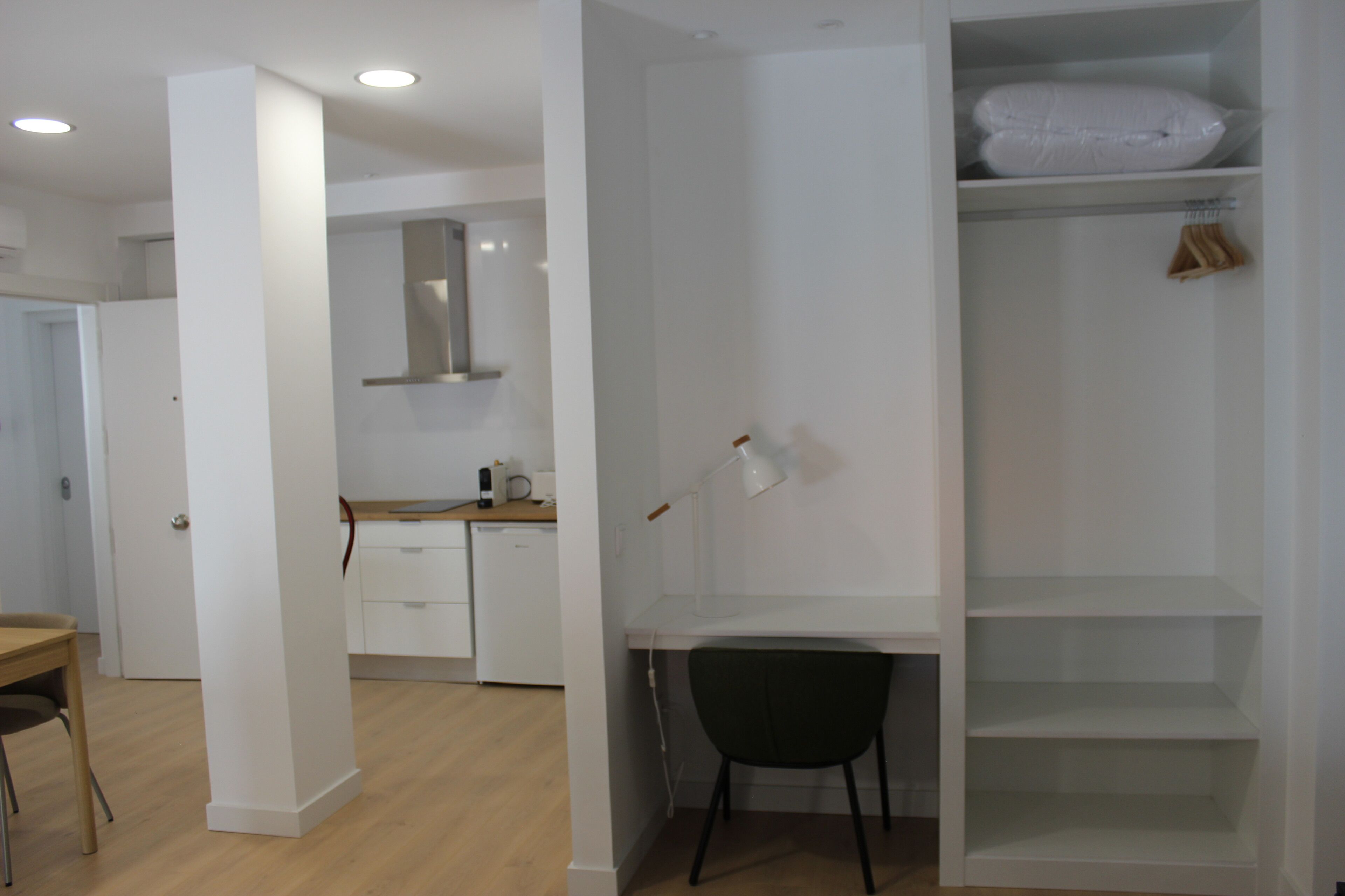 Een moderne, minimalistische kamer met een wit bureau, een stoel en een inbouwgarderobe met planken.