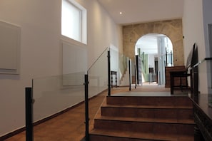 Una escalera con barandillas de vidrio que conduce a un rellano, un muro de piedra y vistas a otra habitación.