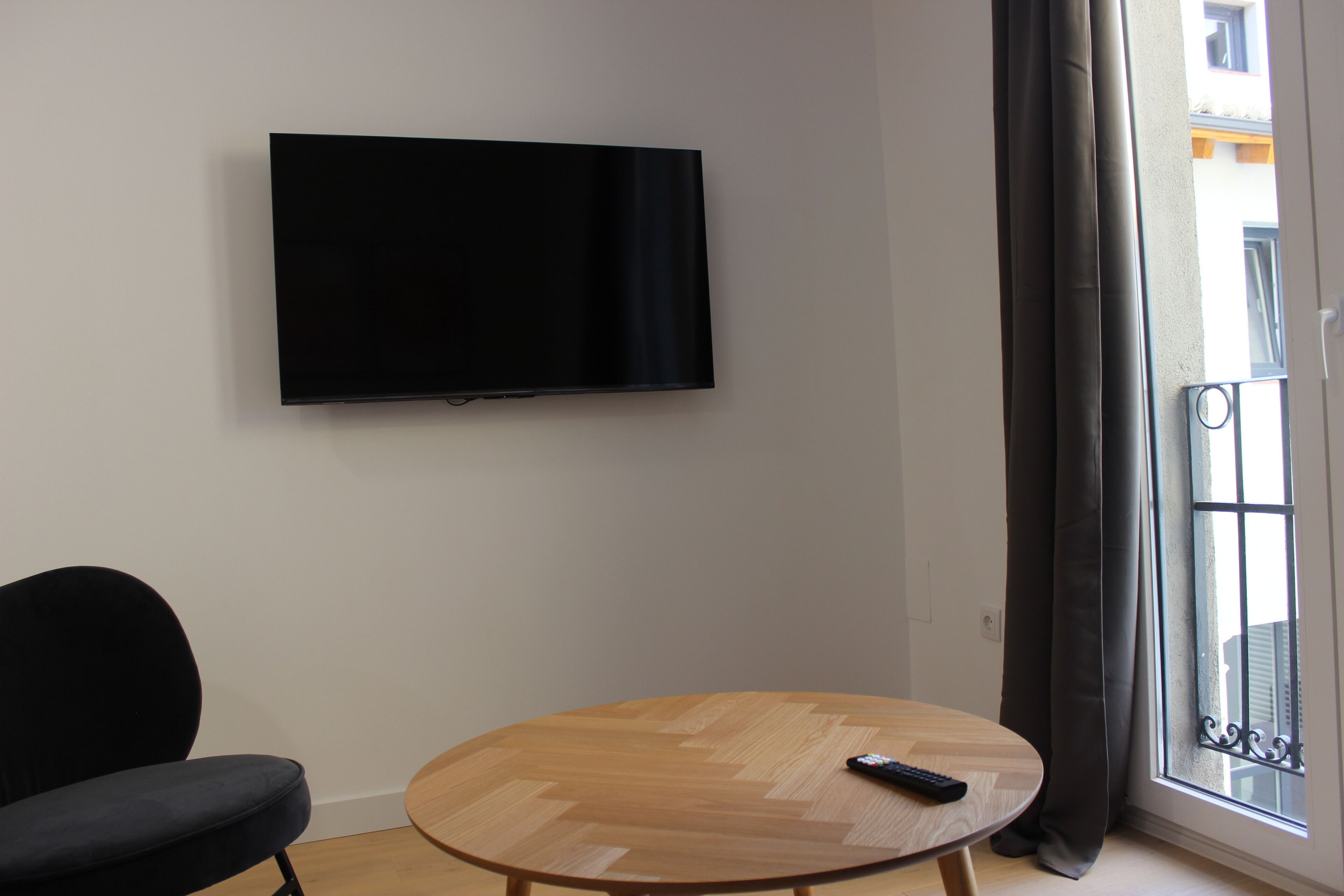 Ein Zimmer mit einem an der Wand befestigten Flachbildfernseher, einem runden Holztisch mit einer Fernbedienung darauf und einem schwarzen Stuhl.