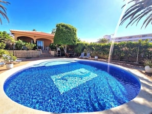 Ein Schwimmbecken mit geometrischem Fliesenmuster, umgeben von einer Terrassenanlage mit Liegestühlen und einem Gebäude im Hintergrund.