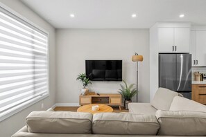 A modern living room with a sofa, a coffee table, a TV, and a kitchen area with cabinets and a refrigerator. - 3 King Beds | Min to UofC | Up and Down Unit (Calgary)