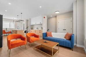 Een moderne woonkamer met een blauwe bank, twee oranje fauteuils, een houten salontafel en een keuken op de achtergrond.