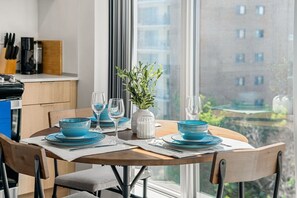Ein Essbereich mit einem runden Tisch für vier Personen, einem Blumentopf und Blick auf ein Gebäude durch ein großes Fenster.
