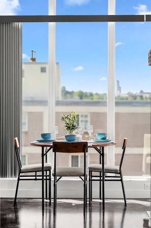 A dining area with a table set for two, a potted plant, and a view of buildings through large windows. - King Bed | UG Park | AC | Mins to Saddledome | 2BR (Calgary)