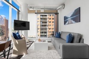 Un salon moderne avec un téléviseur à écran plat, un canapé gris agrémenté de coussins bleus, une table basse blanche et un tapis à motifs.