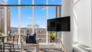 Ett modernt rum med ett stort fönster, en platt-TV på ett stativ, en soffa med en kudde och ett matbord med en vas och ett ljus.