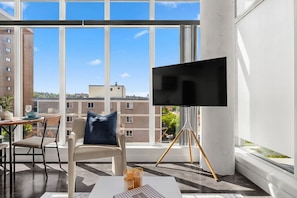 Ein modernes Zimmer mit großem Fenster, einem Flachbildfernseher auf Stativ, einer Couch mit Kissen sowie einem Esstisch mit Vase und Kerze.