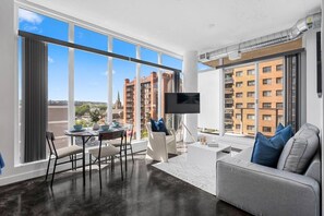 A modern living room with a large window, a flat-screen TV, a sofa, and a dining area. - King Bed | UG Park | AC | Mins to Saddledome | 2BR (Calgary)