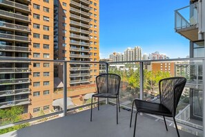 A balcony with two chairs, a glass railing, and a view of high-rise buildings. - King Bed | UG Park | AC | Mins to Saddledome | 2BR (Calgary)