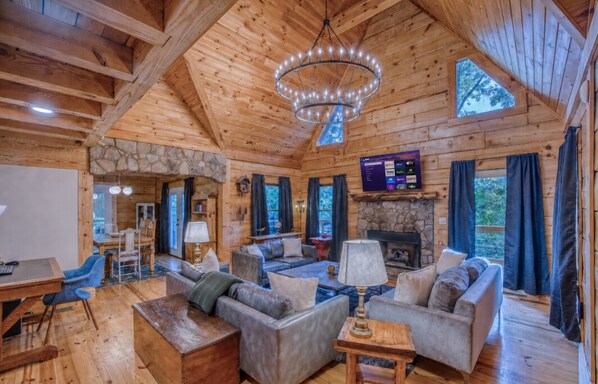 A log cabin interior with a stone fireplace, wooden furniture, and a chandelier.
