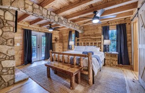 A log cabin bedroom with a stone wall, wooden ceiling beams, a bed, a bench, and a nightstand.