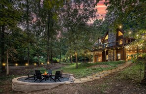 A cabin with a fire pit and outdoor seating, surrounded by trees and a stone staircase leading to the entrance.