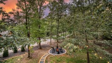 A circular outdoor seating area with a fire pit surrounded by chairs, set in a forested landscape with a clear path leading to it.