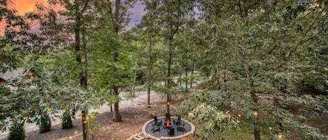 A circular outdoor seating area with a fire pit surrounded by chairs, set in a forested landscape with a clear path leading to it.