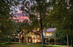 A large, two-story wooden house surrounded by trees with a stone foundation, featuring a covered porch and outdoor lighting.