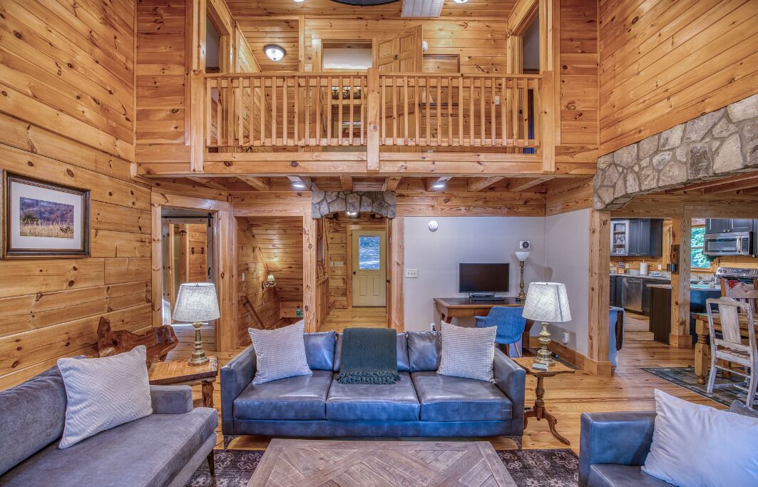 A log cabin interior with a balcony, a living room with a sofa and lamps, and a kitchen area.