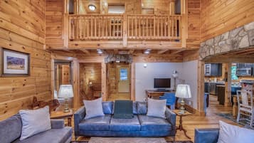 A log cabin interior with a balcony, a living room with a sofa and lamps, and a kitchen area.