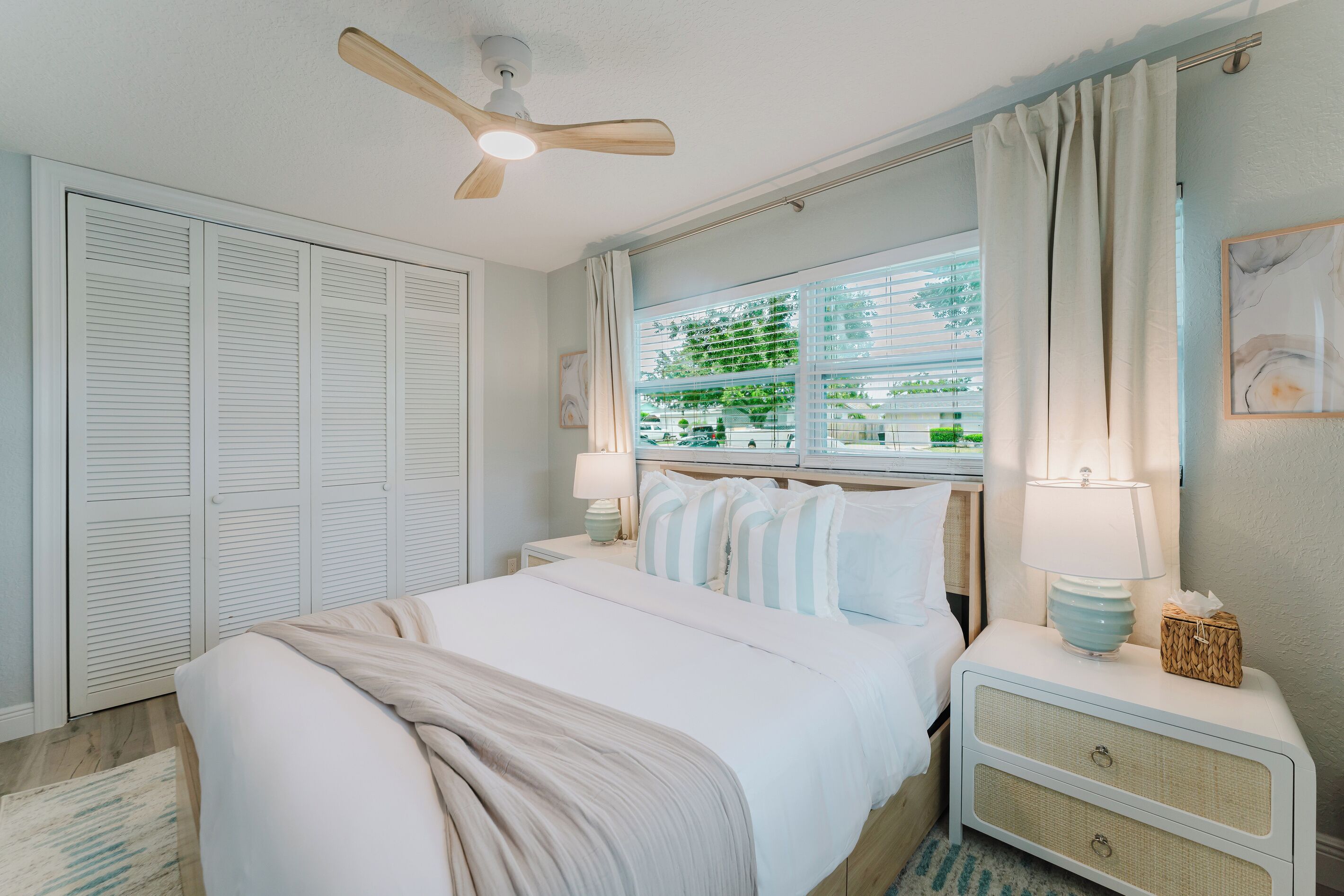 A bedroom with a bed, nightstands, a ceiling fan, a window with curtains, and a view of greenery outside.
