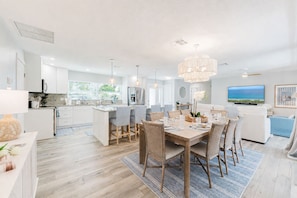 A modern kitchen with white cabinets, a dining area with a wooden table and chairs, and a living room with a sofa and a TV displaying a scenic view. - Designer Pool Oasis | Theater | Golf | Luxury (Palm Beach Gardens)