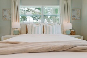 A neatly made bed with a white comforter and striped pillows, flanked by two bedside tables with lamps and decorative items. - Designer Pool Oasis | Theater | Golf | Luxury (Palm Beach Gardens)
