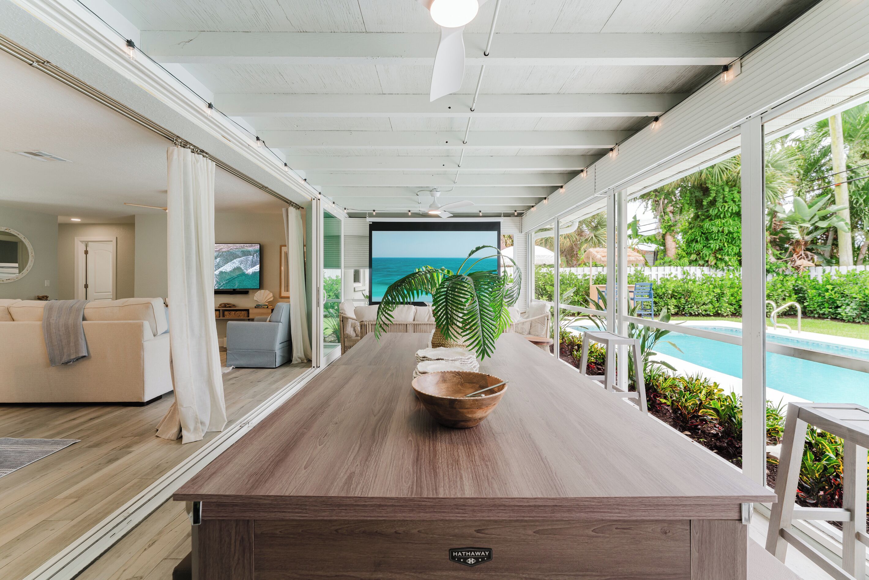 A modern kitchen with a wooden countertop, a large window overlooking a pool and garden, and a television displaying a scenic view.