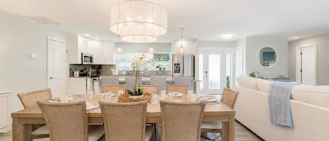 A modern kitchen with white cabinets, a dining area with wicker chairs, and a large chandelier.
