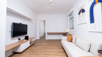 A modern living room with a flat-screen TV, wooden flooring, a white sofa, and a desk with a lamp.