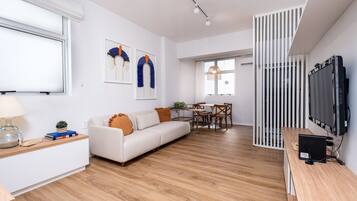 A modern living room with a white sofa, wooden flooring, a dining area with a table and chairs, and a TV mounted on the wall.