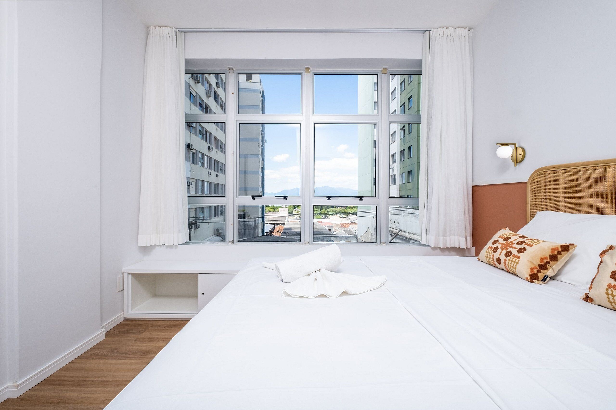 Hotelkamer met een groot raam, een bed met witte beddengoed, een hoofdbord met geweven patroon en een wandlamp.