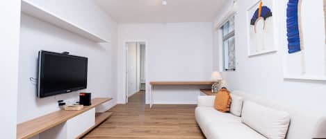 Una sala de estar moderna con una televisión de pantalla plana, suelo de madera, un sofá blanco y un escritorio con una lámpara.