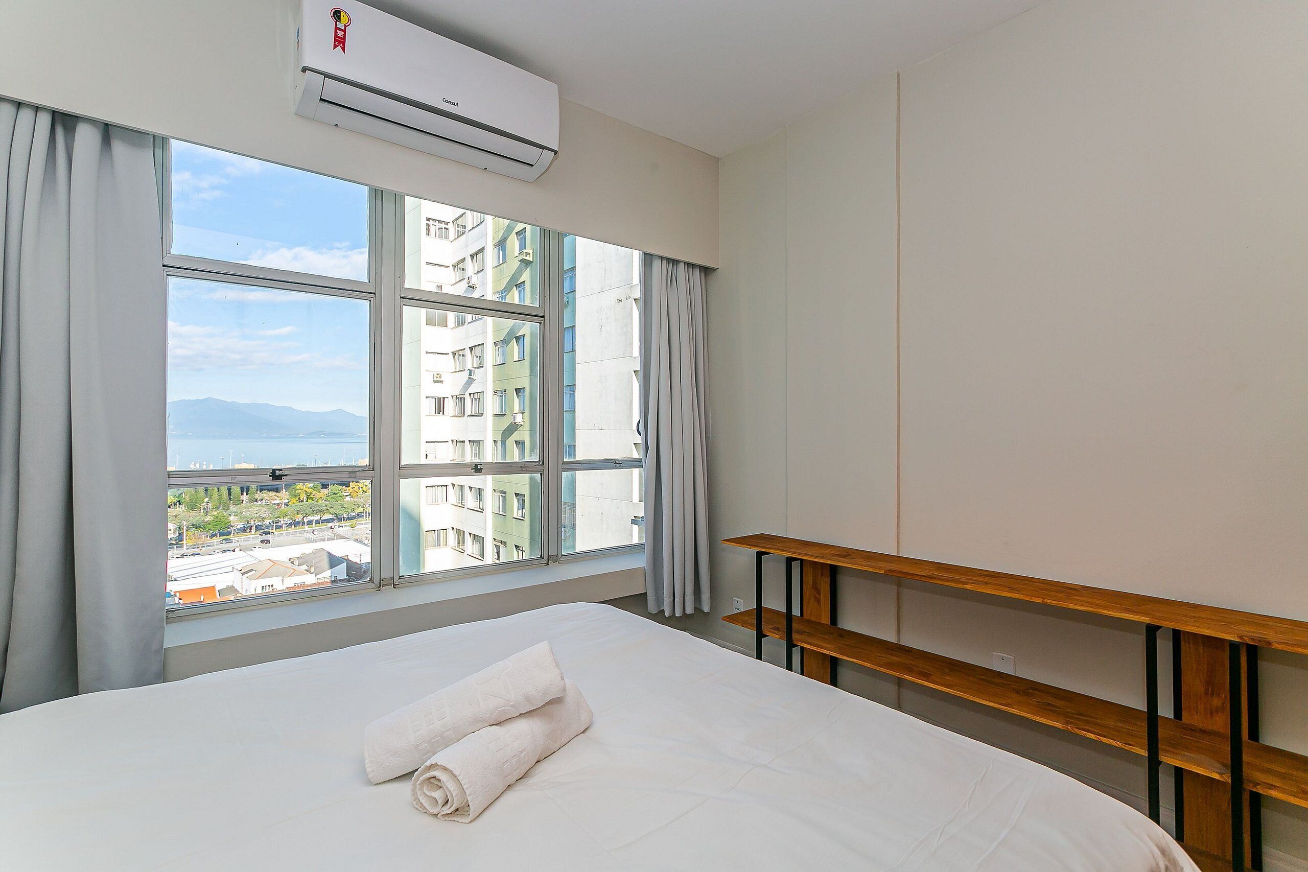 A hotel room with a large bed, a wooden nightstand, and a view of buildings and mountains through the window.