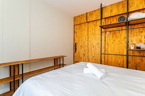 Un lit bien fait, avec une serviette pliée, un banc en bois et une armoire en bois dotée d’étagères.