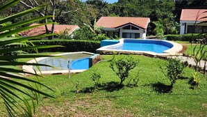 Zona de piscina con una piscina pequeña y otra más grande, rodeada de césped y árboles bien cuidados.