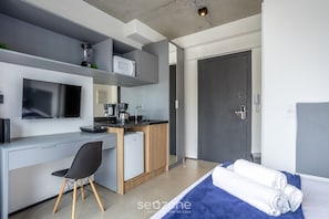 A modern hotel room with a grey and white color scheme, a flat-screen TV, a small kitchenette, a desk with a chair, and a bed with a blue blanket. - Studio w/ Balcony in Bela Vista Vnu1613 (São Paulo)