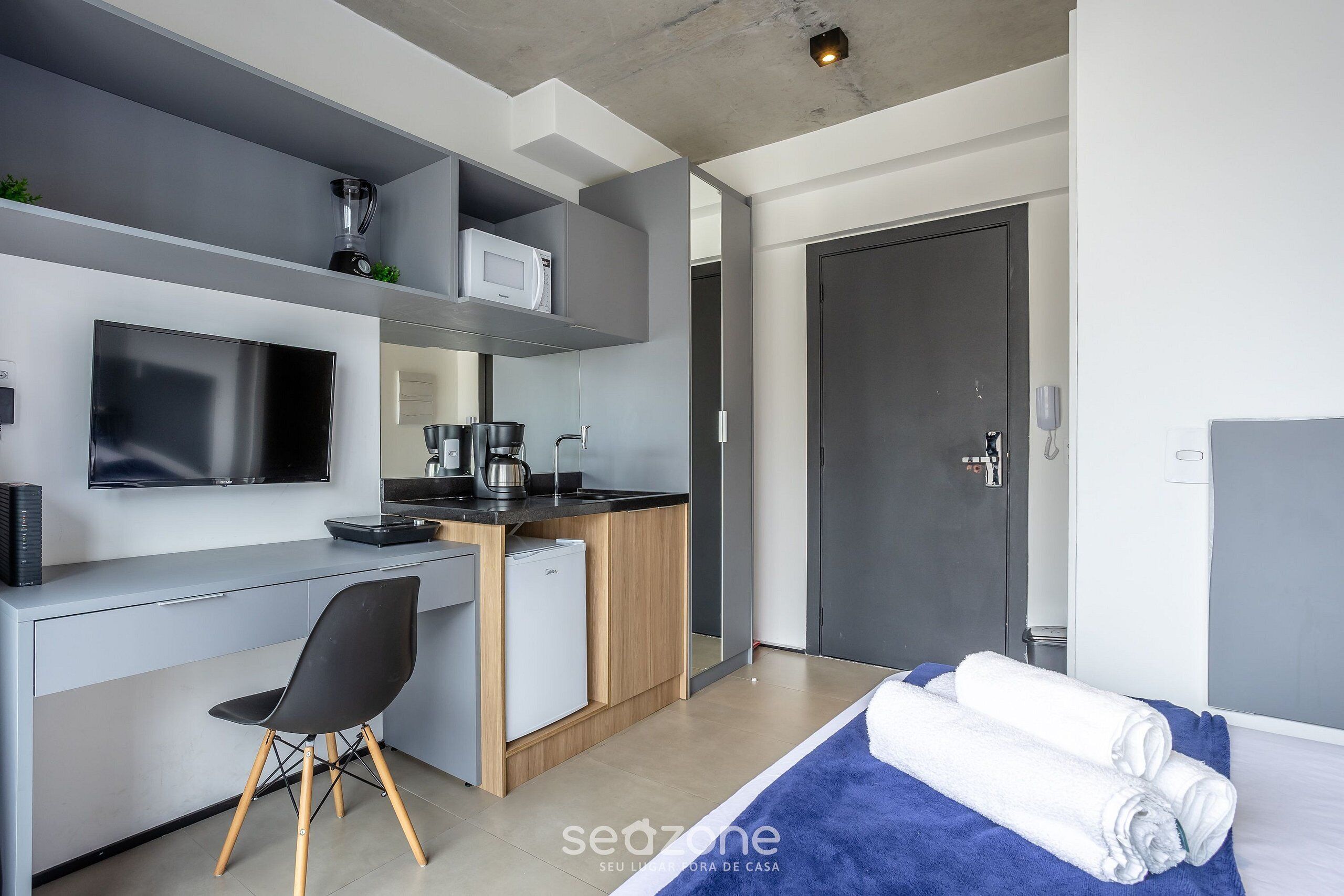 A modern hotel room with a grey and white color scheme, a flat-screen TV, a small kitchenette, a desk with a chair, and a bed with a blue blanket.
