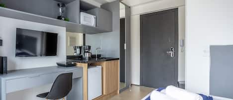 A modern hotel room with a grey and white color scheme, a flat-screen TV, a small kitchenette, a desk with a chair, and a bed with a blue blanket.